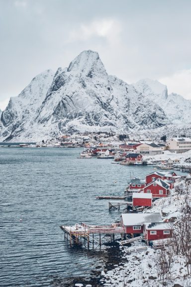 Paisaje invernal con montañas nevadas y cabañas rojas junto a un fiordo.